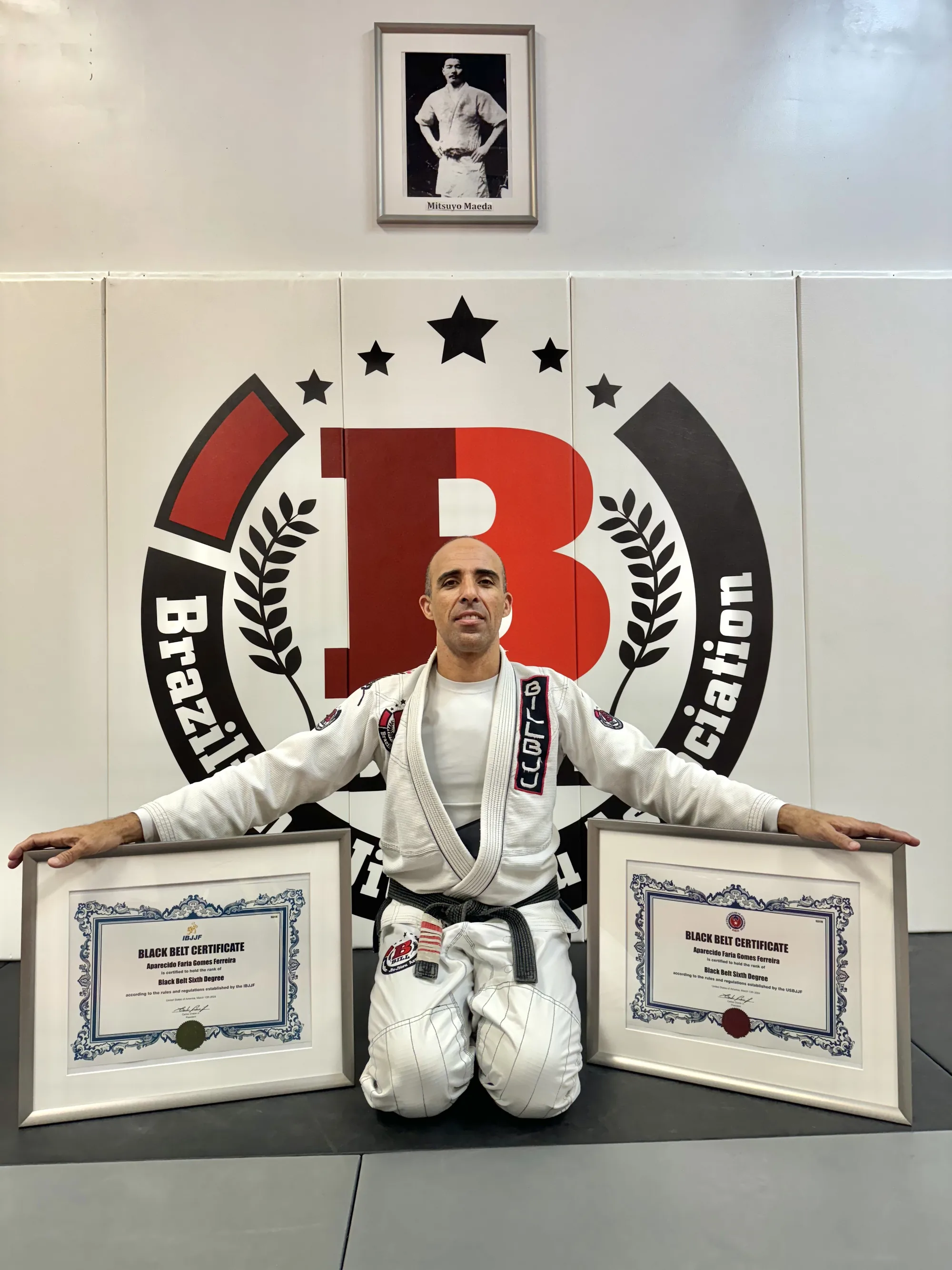 Professor Bill kneeling in front of the academy logo with his two 6th degree black belt certificates