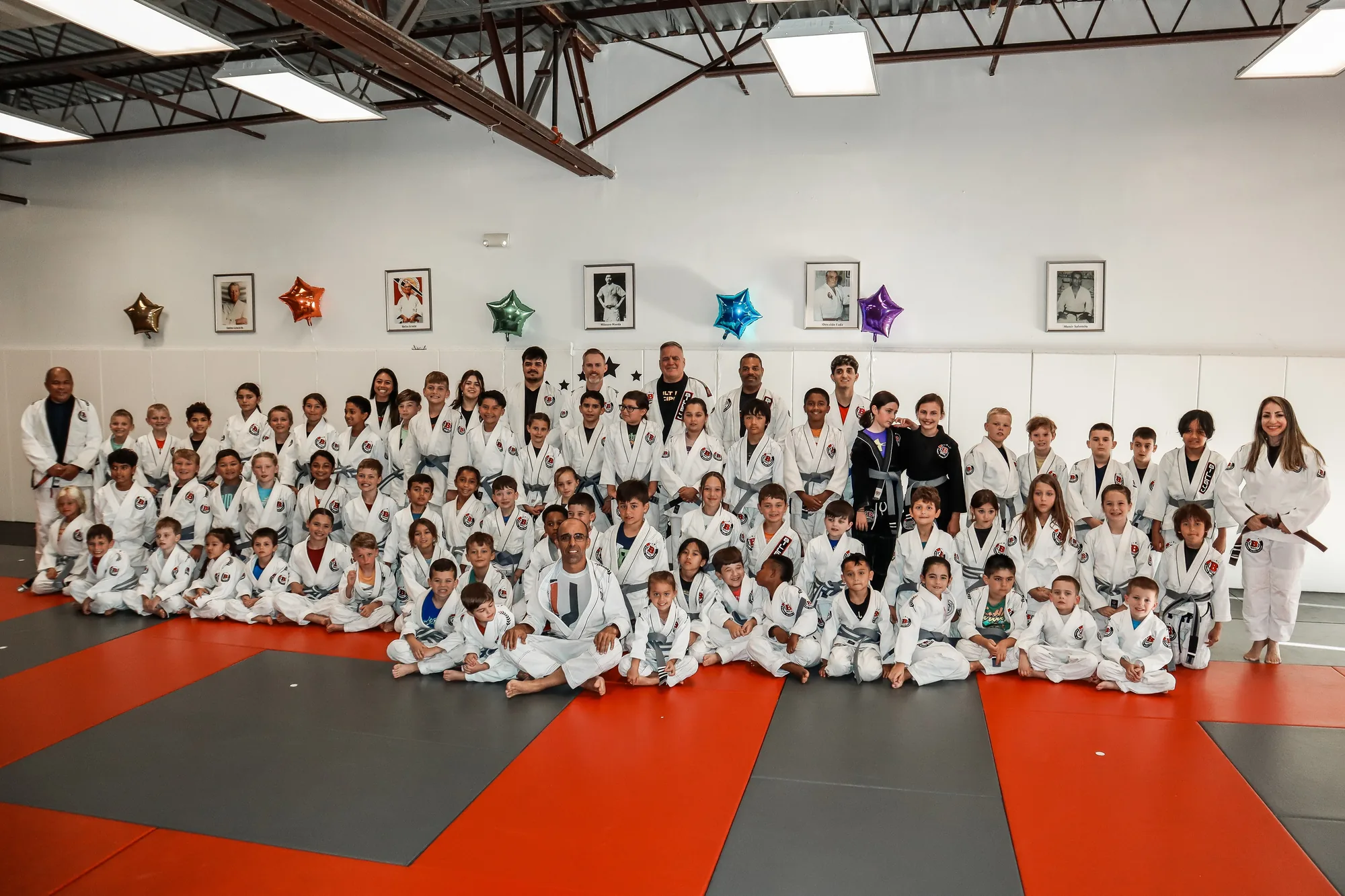 Professor Bill sitting in the middle of his kids class, dozens of students in white gis on the red and gray mats