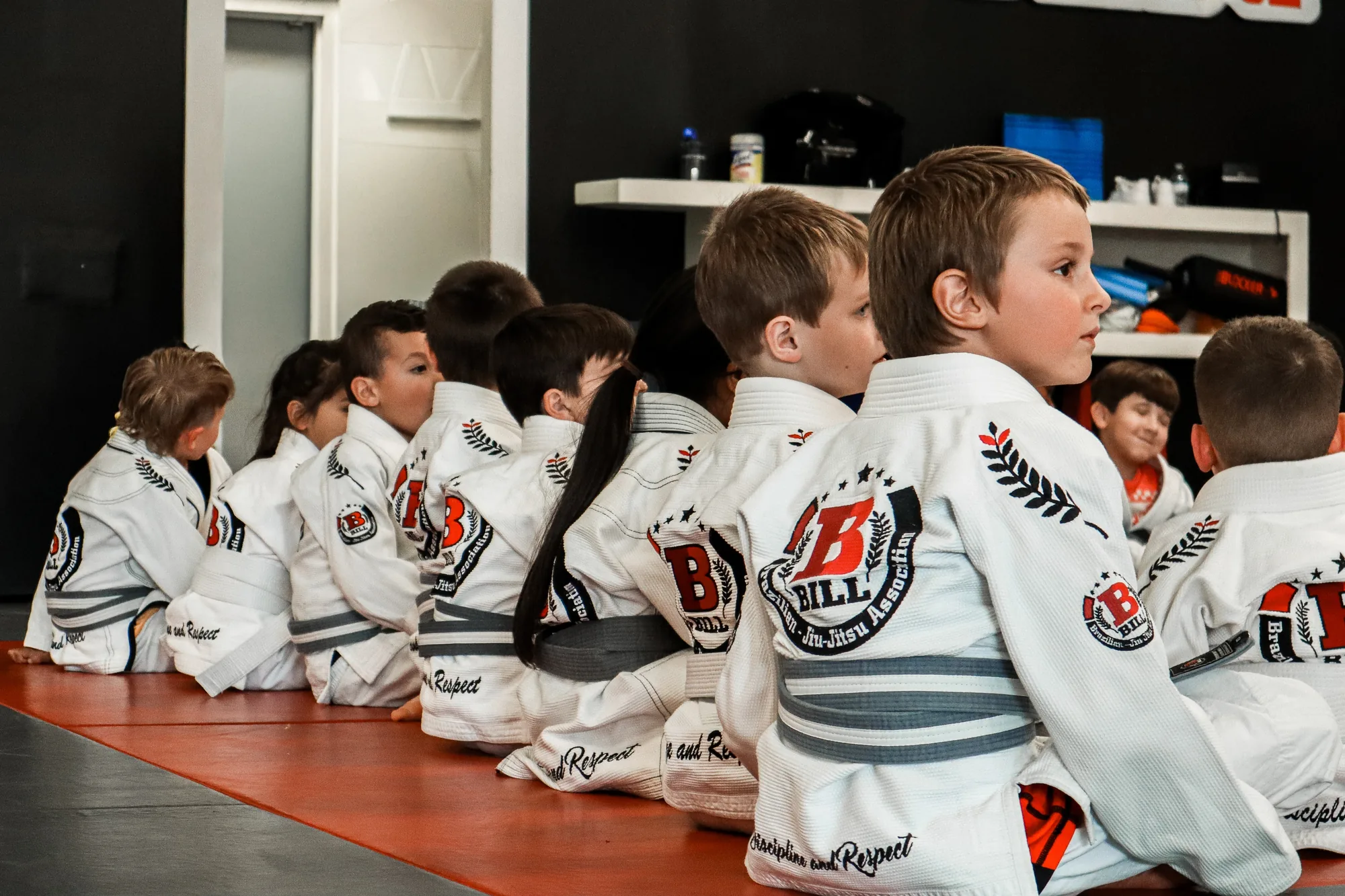 Young students in white gis seated in a line on the mat, one boy turning to look ahead