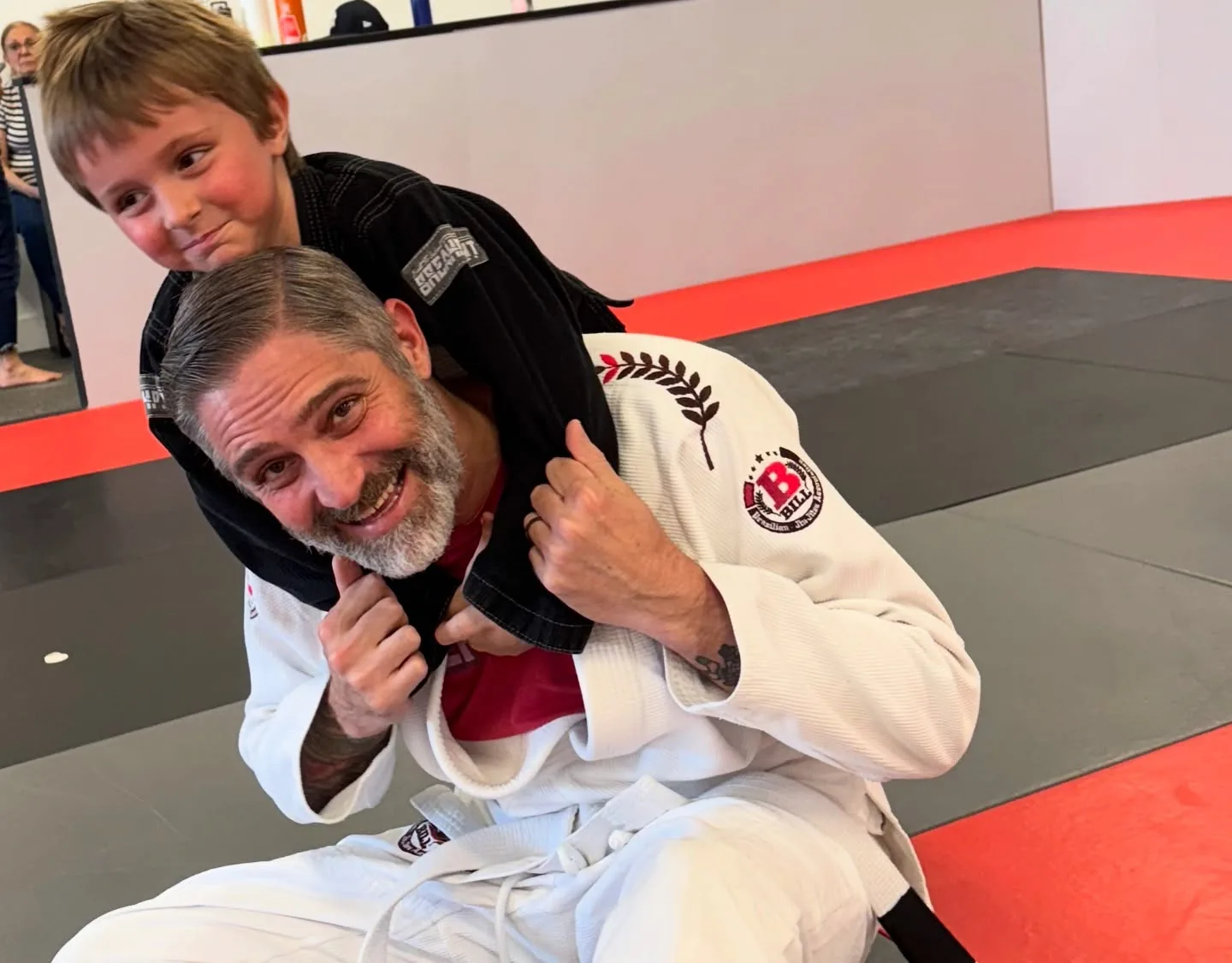 A smiling dad in a white gi with his son hanging on his back on the mat